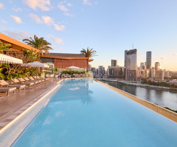 Rooftop infinity pool with lounge chairs, umbrellas, and palm trees overlooking a modern city skyline at sunrise or sunset.