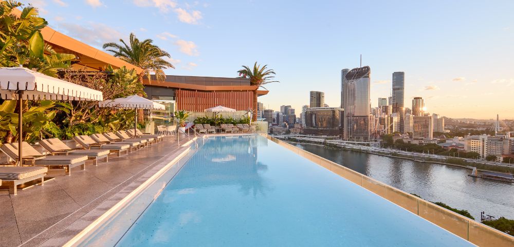 Rooftop infinity pool with lounge chairs, umbrellas, and palm trees overlooking a modern city skyline at sunrise or sunset.