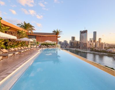 Rooftop infinity pool with lounge chairs, umbrellas, and palm trees overlooking a modern city skyline at sunrise or sunset.