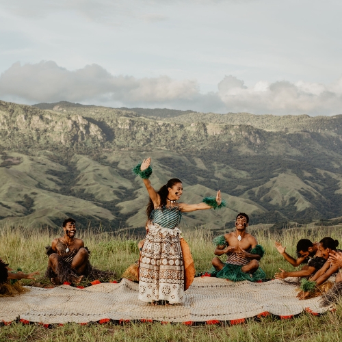 A woman performs a traditional dance on a woven mat outdoors, surrounded by sitting participants. They wear cultural attire; distant mountains form the backdrop.