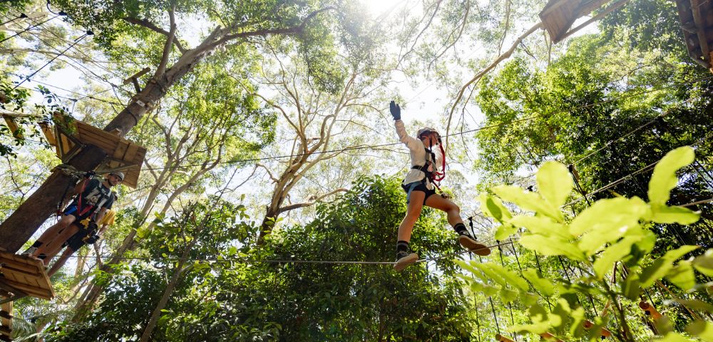 A person wearing safety gear and a harness navigating a high ropes obstacle in a forest treetop adventure course.