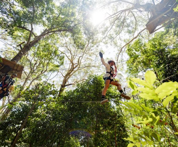 A person wearing safety gear and a harness navigating a high ropes obstacle in a forest treetop adventure course.
