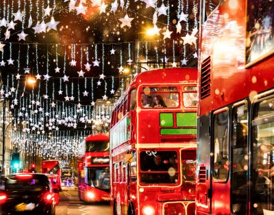 A busy London street features double-decker buses and glowing holiday decorations at night.