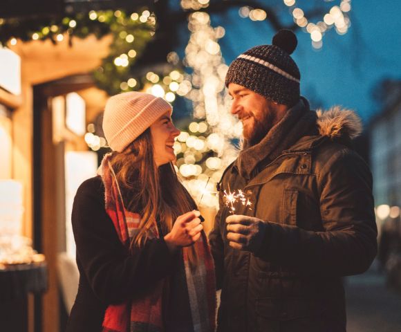 A couple shares a moment beneath twinkling festive lights at a holiday market.