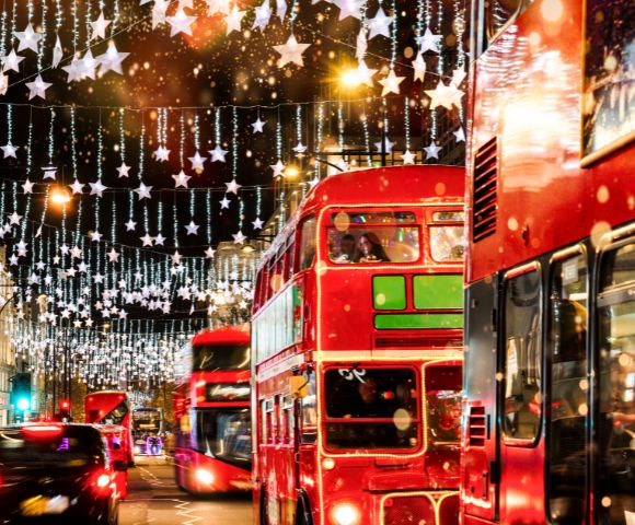 A busy London street features double-decker buses and glowing holiday decorations at night.