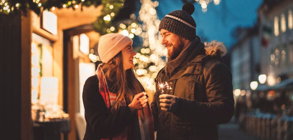 A couple shares a moment beneath twinkling festive lights at a holiday market.