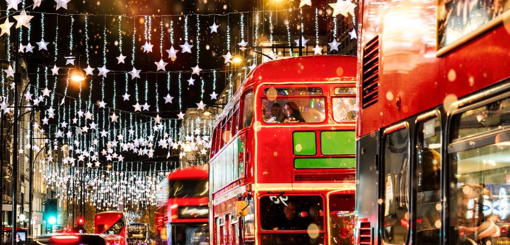 A busy London street features double-decker buses and glowing holiday decorations at night.