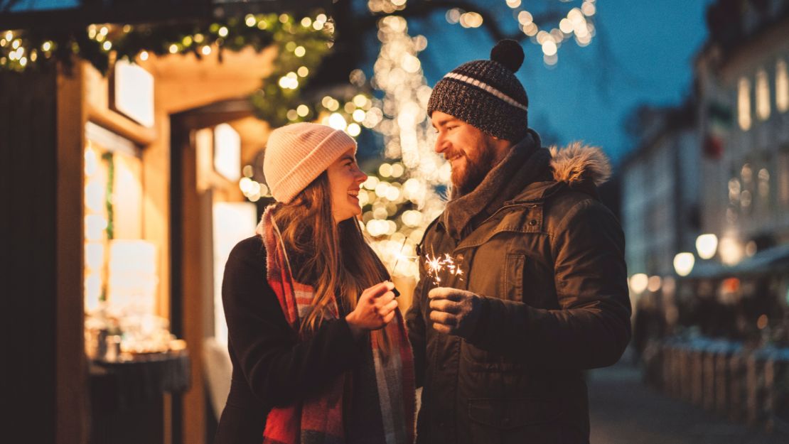 A couple shares a moment beneath twinkling festive lights at a holiday market.