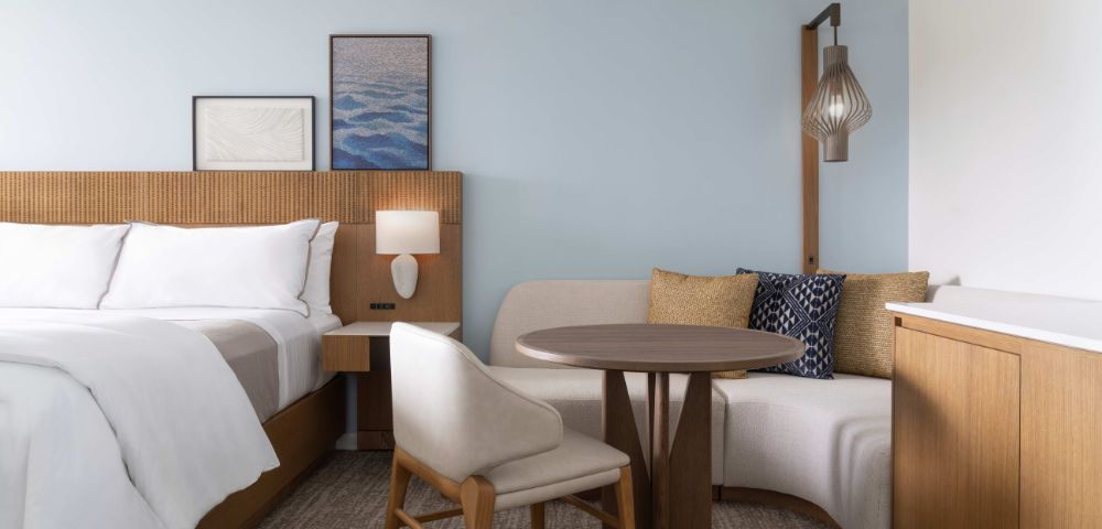 Hotel room with a neatly made bed, wooden headboard, framed artwork, a round table with two chairs, and a cushioned bench under a hanging light fixture.