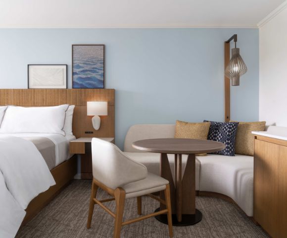 Hotel room with a neatly made bed, wooden headboard, framed artwork, a round table with two chairs, and a cushioned bench under a hanging light fixture.