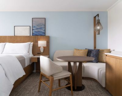 Hotel room with a neatly made bed, wooden headboard, framed artwork, a round table with two chairs, and a cushioned bench under a hanging light fixture.