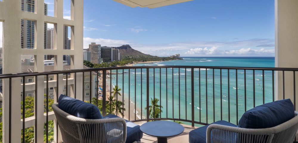 Balcony with two blue-cushioned chairs and a small round table, overlooking the beach, ocean, nearby buildings, and distant mountains.