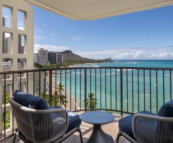 Balcony with two blue-cushioned chairs and a small round table, overlooking the beach, ocean, nearby buildings, and distant mountains.