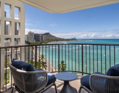Balcony with two blue-cushioned chairs and a small round table, overlooking the beach, ocean, nearby buildings, and distant mountains.