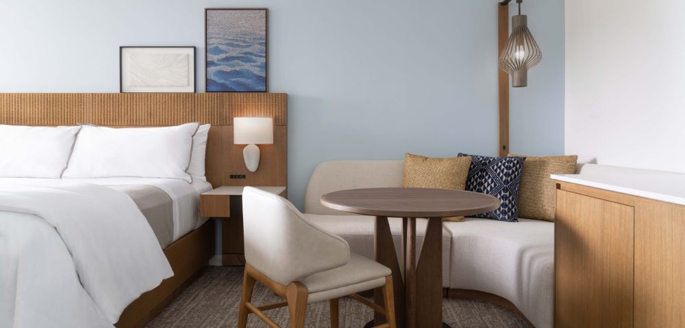 Modern hotel room with a neatly made bed, white linens, round table with two chairs, framed artwork, and a bedside lamp