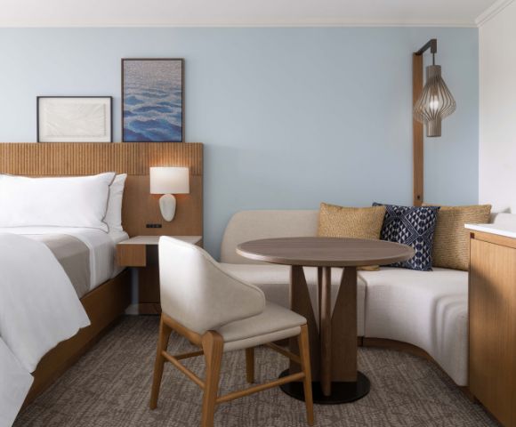 Modern hotel room with a neatly made bed, white linens, round table with two chairs, framed artwork, and a bedside lamp