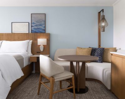 Modern hotel room with a neatly made bed, white linens, round table with two chairs, framed artwork, and a bedside lamp