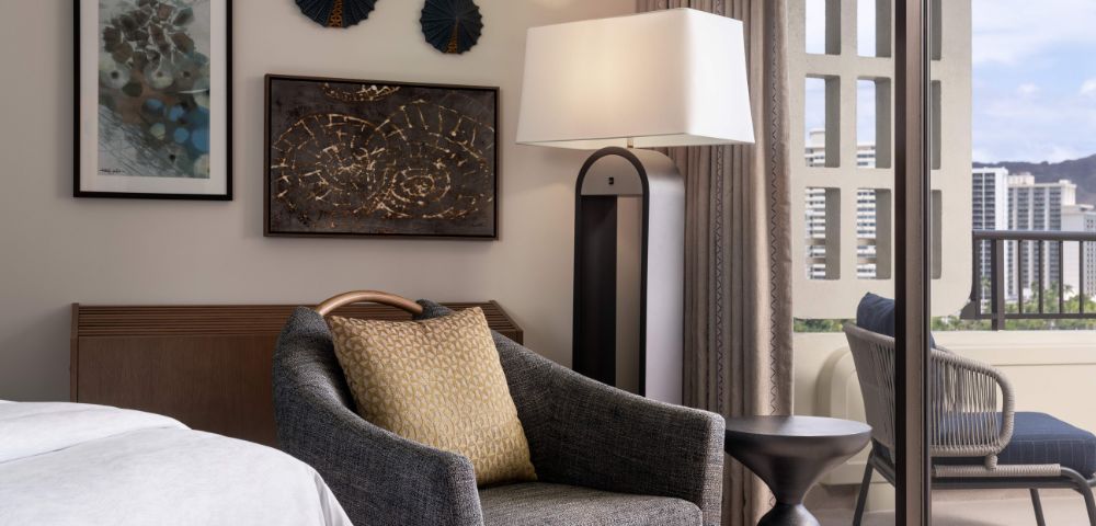 Hotel room corner with a bed, dark armchair with a light brown cushion, wooden nightstand with a lamp, framed artwork, and curtains over large windows.