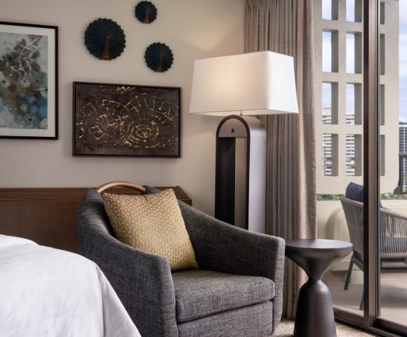 Hotel room corner with a bed, dark armchair with a light brown cushion, wooden nightstand with a lamp, framed artwork, and curtains over large windows.