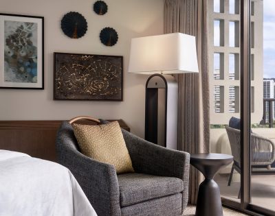Hotel room corner with a bed, dark armchair with a light brown cushion, wooden nightstand with a lamp, framed artwork, and curtains over large windows.