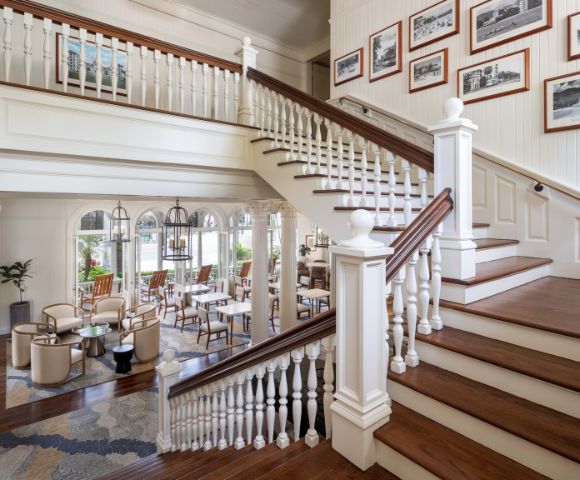 Grand staircase with white railings and wooden steps, illuminated by natural light from large windows, with seating visible in the background.