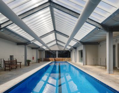 Indoor swimming pool with a glass roof, surrounded by seating areas and columns, illuminated by natural light.