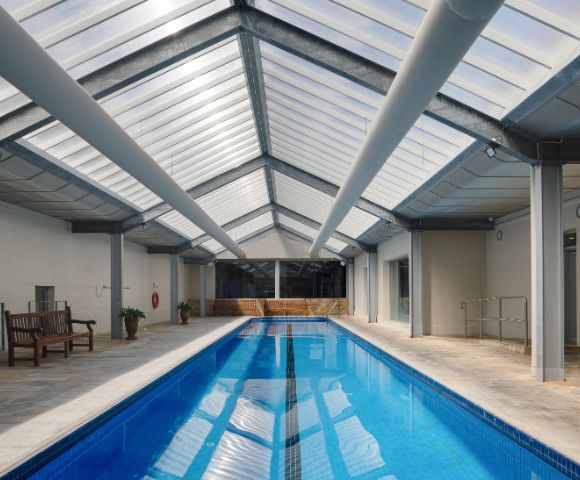 Indoor swimming pool with a glass roof, surrounded by seating areas and columns, illuminated by natural light.