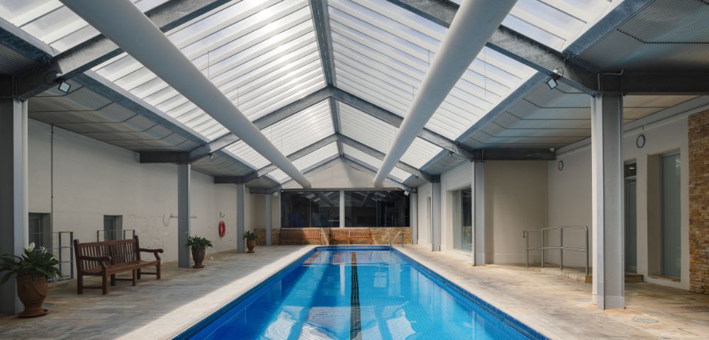 Indoor swimming pool with a glass roof, surrounded by seating areas and columns, illuminated by natural light.