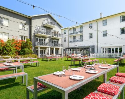 Outdoor dining area in front of a hotel or large building, with tables set for dining and greenery surrounding the space.