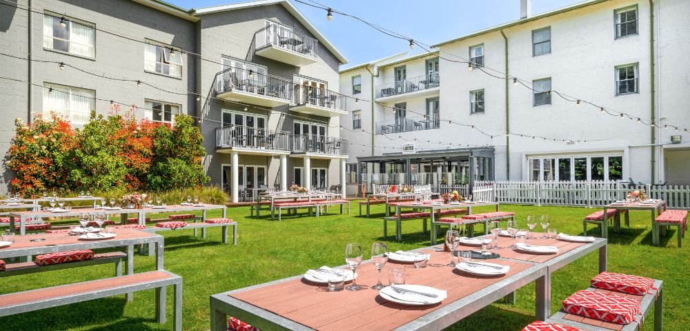 Outdoor dining area in front of a hotel or large building, with tables set for dining and greenery surrounding the space.