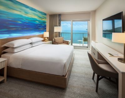 Alternate angle of the hotel room showing two beds, a seating area, and a desk near large windows with ocean scenery.