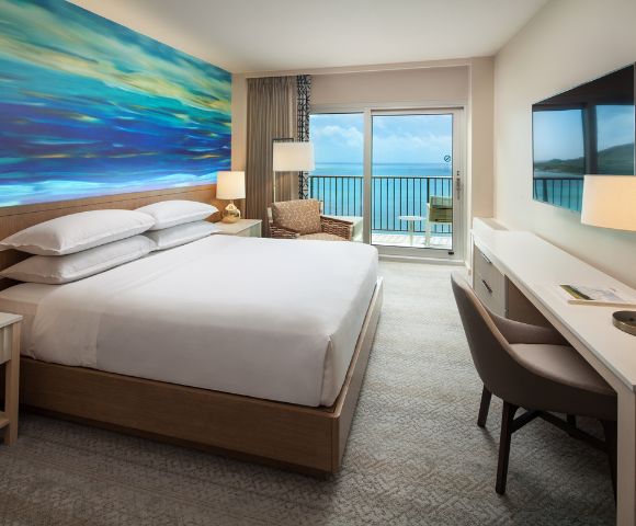 Alternate angle of the hotel room showing two beds, a seating area, and a desk near large windows with ocean scenery.