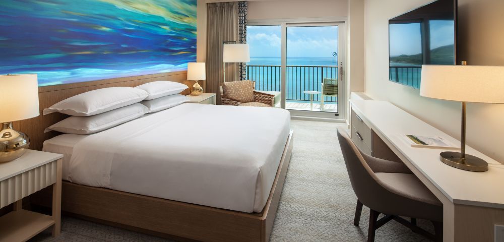 Alternate angle of the hotel room showing two beds, a seating area, and a desk near large windows with ocean scenery.