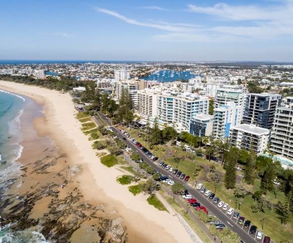 Aerial view of a long sandy beach with waves crashing on the shore, bordered by tall buildings and greenery.