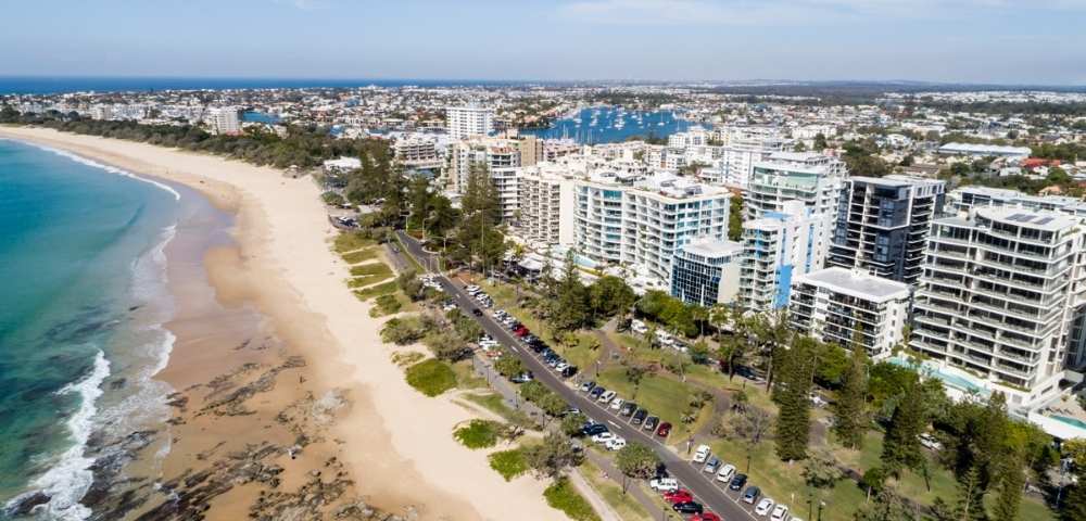 Aerial view of a long sandy beach with waves crashing on the shore, bordered by tall buildings and greenery.