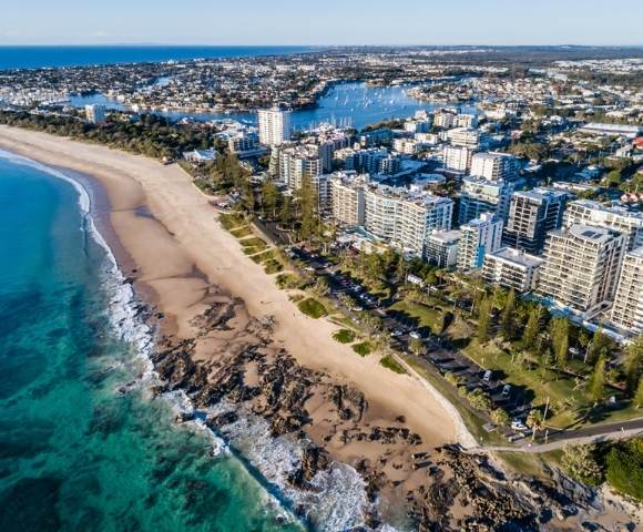 Aerial view of a sandy beach with clear blue water and numerous buildings along the coast.