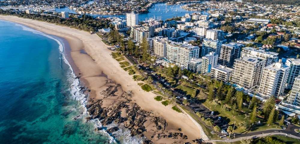 Aerial view of a sandy beach with clear blue water and numerous buildings along the coast.