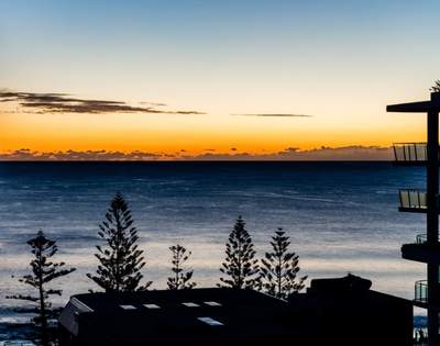 Scenic sunset view over the ocean with silhouettes of trees and buildings in the foreground.