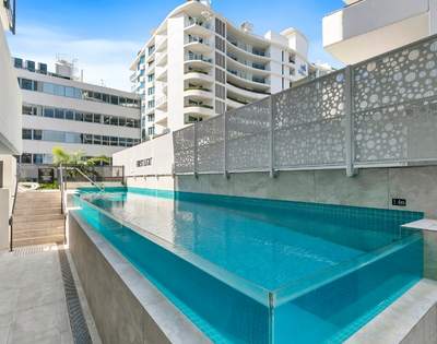 Outdoor swimming pool in a modern apartment complex, surrounded by buildings and enclosed with glass fencing.