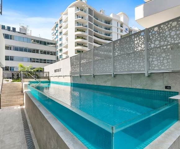 Outdoor swimming pool in a modern apartment complex, surrounded by buildings and enclosed with glass fencing.