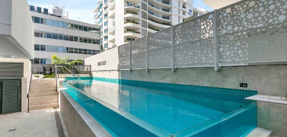 Outdoor swimming pool in a modern apartment complex, surrounded by buildings and enclosed with glass fencing.