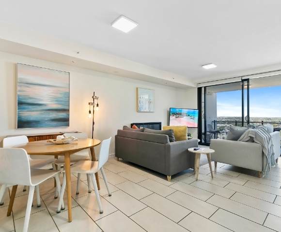 Open-plan living area with a dining table, chairs, sofa set, TV, and large windows providing an expansive outdoor view.