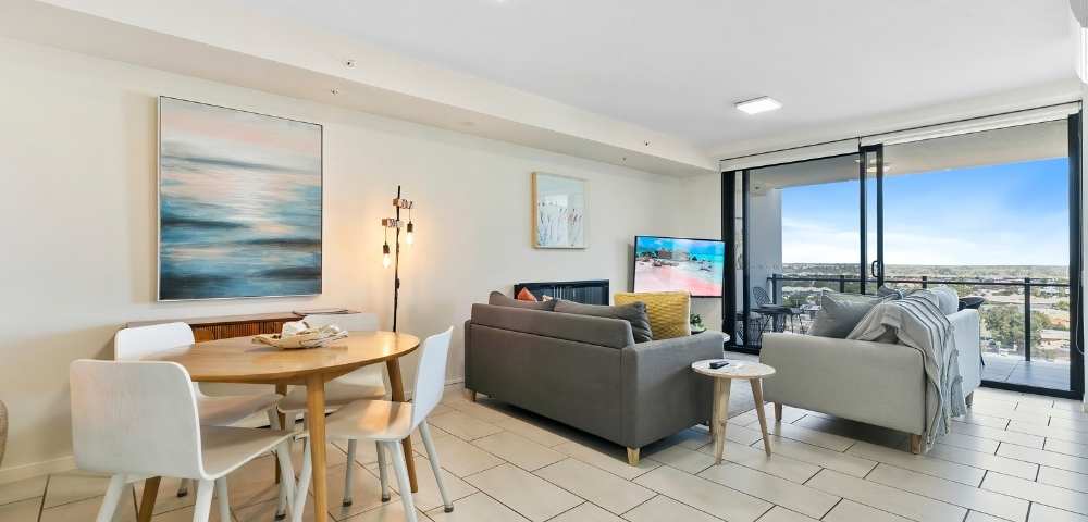 Open-plan living area with a dining table, chairs, sofa set, TV, and large windows providing an expansive outdoor view.