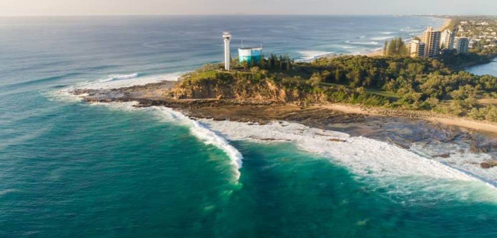 Aerial shot of a rocky coastline with waves crashing and a lighthouse visible in the distance.