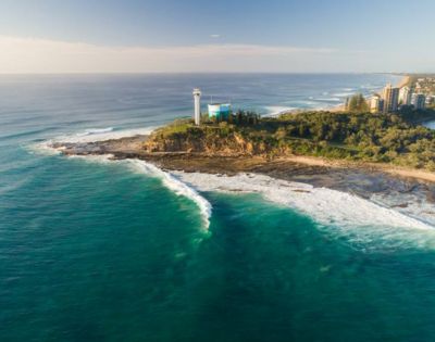Aerial shot of a rocky coastline with waves crashing and a lighthouse visible in the distance.