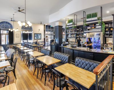 Cozy restaurant or café interior with wooden tables and chairs, a bar counter, and shelves stocked with bottles.