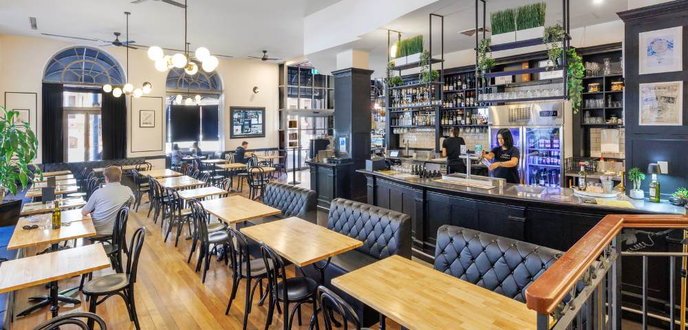 Cozy restaurant or café interior with wooden tables and chairs, a bar counter, and shelves stocked with bottles.