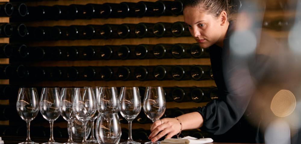 Person arranging multiple empty wine glasses on a table, likely preparing for a wine tasting session.