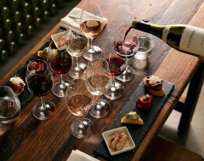 Table set with multiple wine glasses filled with different wines, a bottle being poured, and small plates with food items, depicting a wine tasting experience.