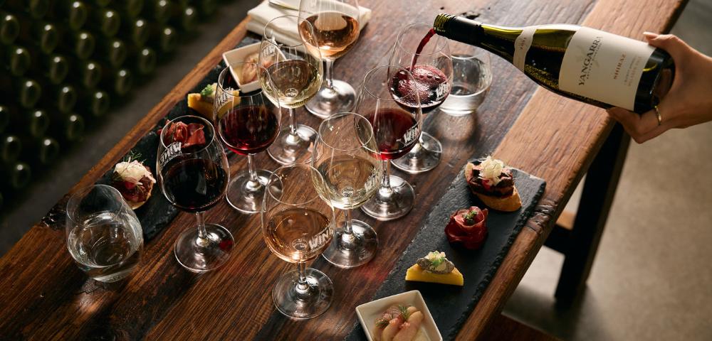 Table set with multiple wine glasses filled with different wines, a bottle being poured, and small plates with food items, depicting a wine tasting experience.